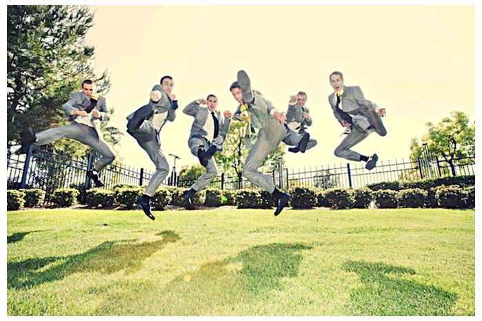 Casual groomsmen jump, ninja style, in green open field for this fun wedding day action shot