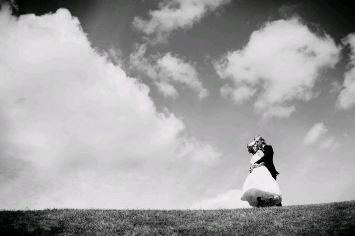 Bride and groom atop a hill, hold each other with a beautiful cloudy sky in the background