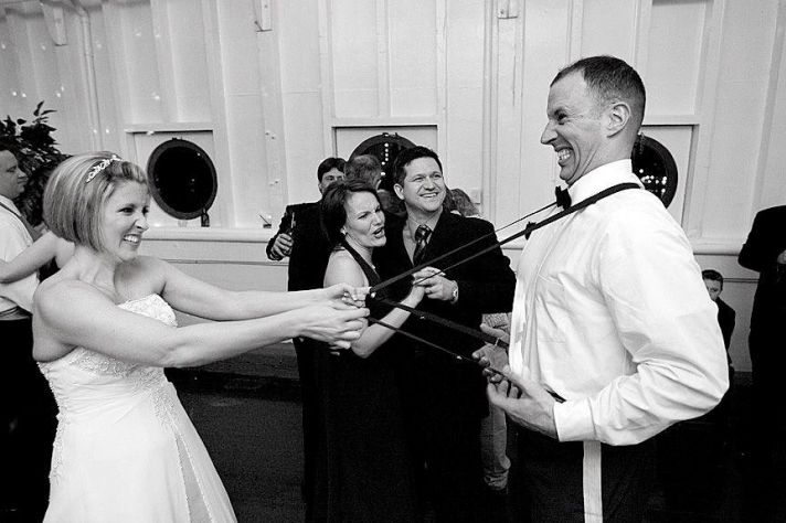 Black and white photo of bride having fun with groom’s suspenders at wedding reception