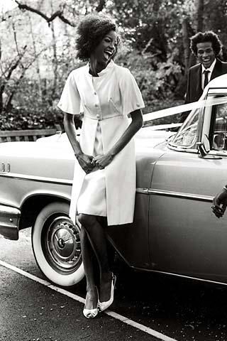 Beautiful bride-to-be leans against antique car, wearing white short sleeve coat dress