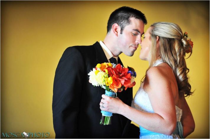 Bride in lace halter wedding dress, holds vibrant bridal bouquet, kisses groom in traditional black 