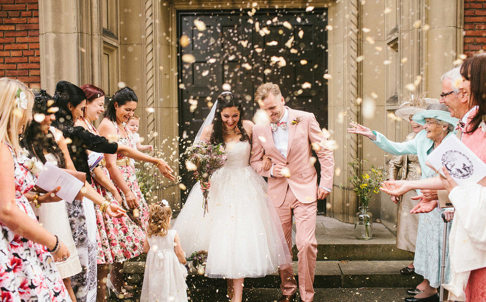 Groom exits ceremony church with his bride wearing pale coral suit and bow tie